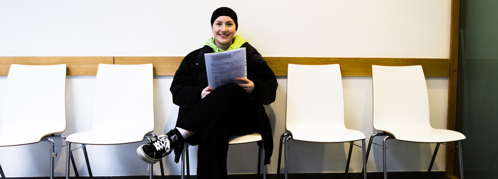 Junge Frau, die in einem Wartebereich auf einem Stuhl sitzt und Dokumente in den Händen hält.